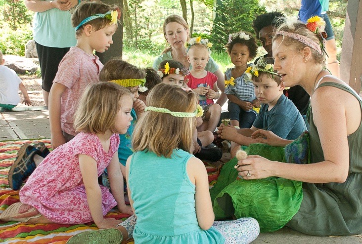 A group of toddlers and parents sit in a circle outdoors and look on as a teacher engages the toddlers through their imagination by telling a story using a puppet.