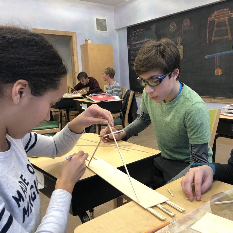A middle school girl and boy in the foreground work on a physics experiment, while two boys in the distant background also work on a science experiment. 