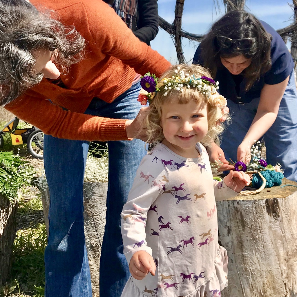 Mom adjusts flower crown on smiling toddler at May Fair, while parent in background works on making more crowns. 
