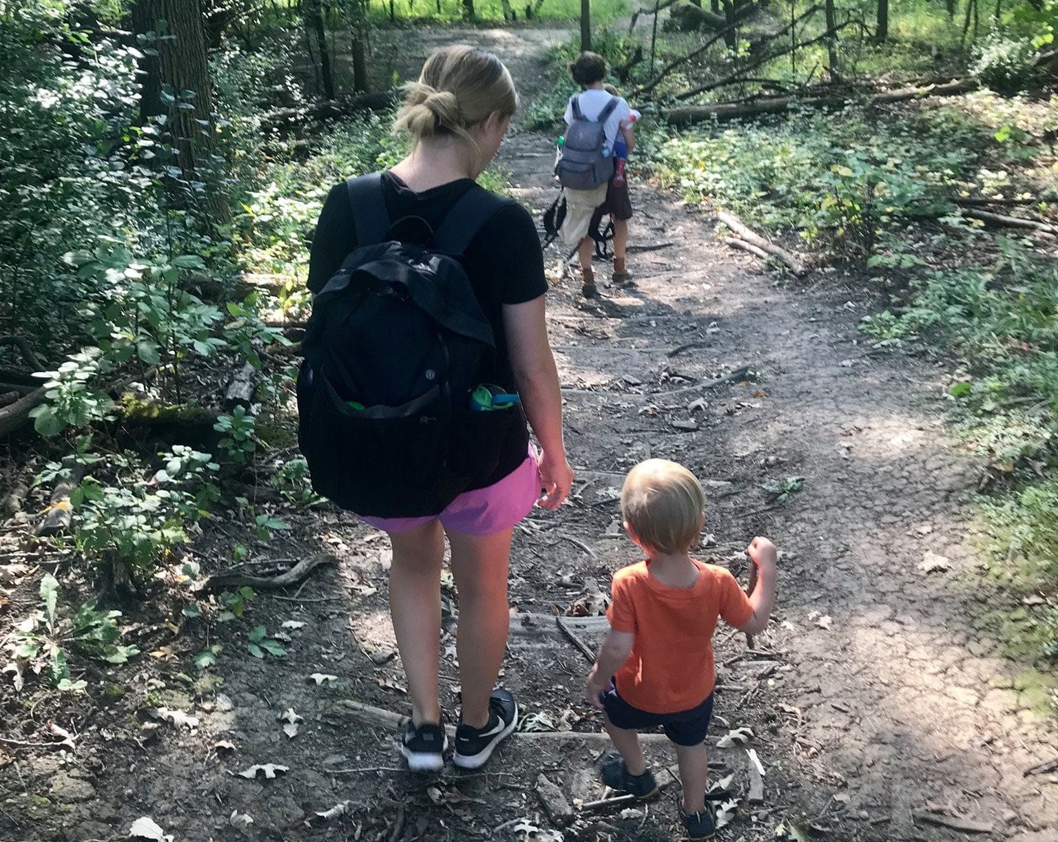 A mother and child in the foreground and a mother and child in the background hike down a woodland trail as part of nature exploration in our Child in the Wild Playgroup.